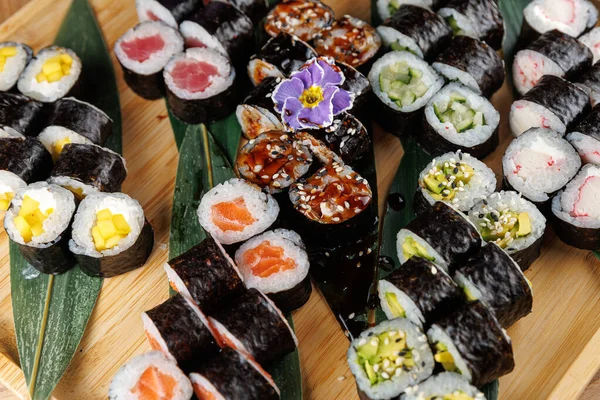Çeşitli suşi ruloları yeşil yapraklı ahşap bir tabakta düzenlenir. Ekmekler çeşitli dolgular ve malzemeler içeriyor. Japon mutfak sanatı sergileniyor..
