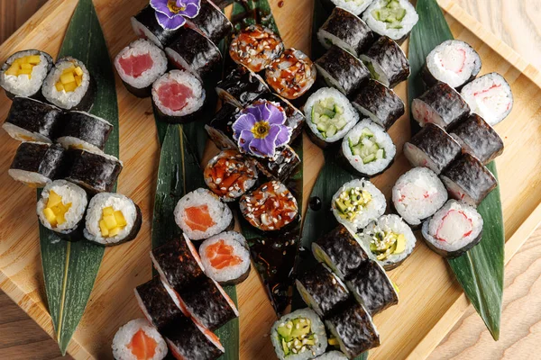 Tabaklarda farklı deniz ürünleri ve sebzelerden yapılan renkli suşi ruloları ahşap bir tahtada güzelce sıralanmış. Dekoratif yapraklar sunumu güçlendirir.