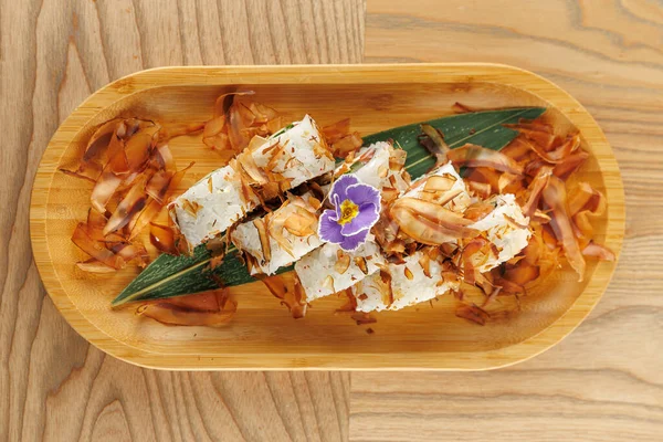 Ahşap tepside güzel düzenlenmiş suşi var. Üstünde narin bonito taneleri ve mor bir çiçekle süslenmiş. Ayar gündelik ve davetkar..