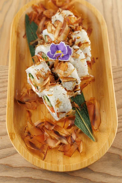 Suşi ruloları bambu tabağa konur. Kepekli malzemeler ve mor yenilebilir bir çiçek sunuma detay ekler. Bu sahne Japon mutfak sanatının bir karışımını gösteriyor..