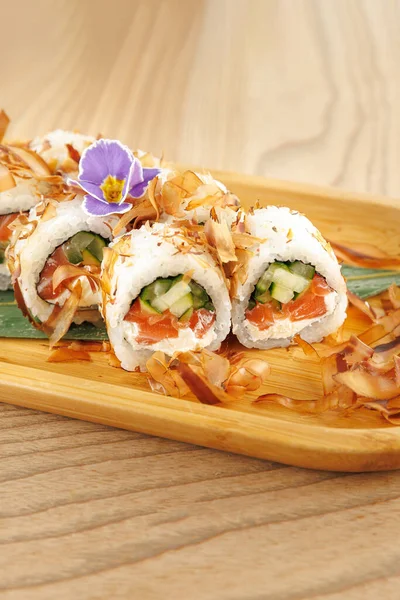 Taze suşi ruloları ahşap bir tabakta güzelce düzenlenir. Ekmeğin içinde salatalık ve somon var. Üstünde bonito gevreği ve çiçekle süslenmiş..