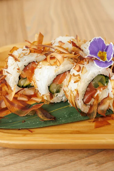 Suşi ruloları ahşap bir tabakta sunuluyor. İçinde balık, salatalık ve çıtır çıtır elementler var. Mor bir çiçek yemeği süslüyor, eşsiz bir dokunuş ekliyor..