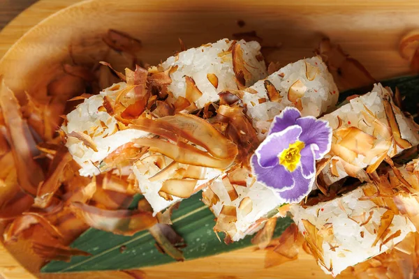 Suşi ruloları ahşap bir tabakta servis edilir. Üstünde bonito taneleri sergilenir. Küçük mor bir çiçek tabağa dekoratif bir dokunuş ekler..