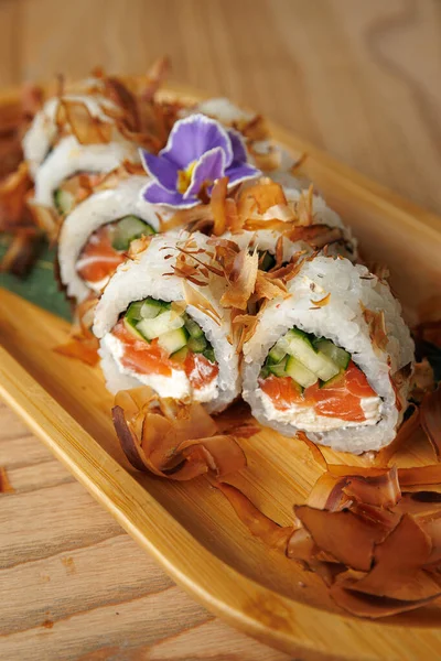 Somon, salatalık ve krem peynirli bambu tabakta lezzetli bir suşi rulosu sunulur. Üstünde çıtır balık pulları ve süsleme için bir çiçek var..