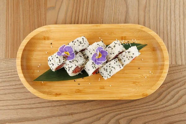 Balık ve susam tohumlu pirinçli suşi ruloları ahşap bir tepside düzenlenir. Yenilebilir çiçekler biraz renk katar. Bu yemek sıradan bir yemek salonunda servis edilir..