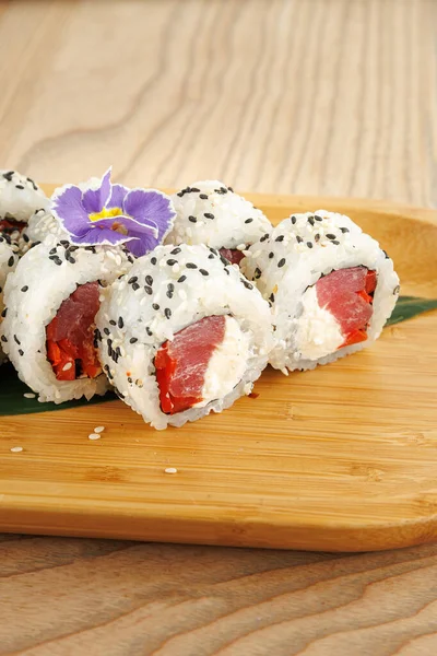 Suşi ruloları ahşap bir tabakta güzelce düzenlenir. Her rulonun yanında pirinç, susam tohumu, ton balığı ve bir çiçek var. Bu yemek Japon yemeklerini temsil ediyor..