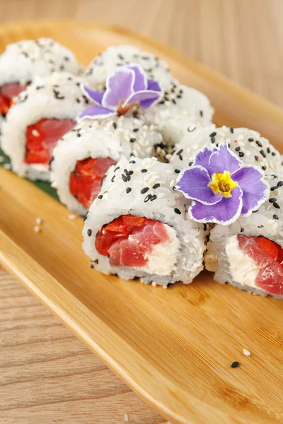 Taze suşi ruloları, mor çiçekler ve susam tohumlarıyla kaplı somon ve krem peynir içerir. Bir bambu tabağında güzelce dizilmişler, eğlenmeye hazırlar..