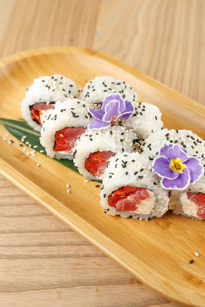 Somon ve krem peynirli suşi ruloları ahşap bir tabakta güzelce düzenlenir. Küçük bir dekoratif çiçek ve susam tohumları sunuma detay ekler.