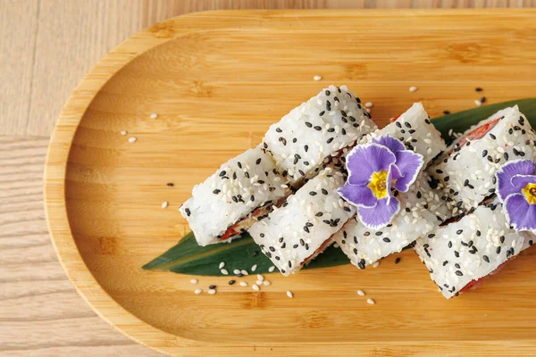 Birkaç suşi rulosu ahşap bir tabakta susam tohumları serpiştirilir ve üzerinde bir çiçek vardır. Tabakta Japon aşçılık ve sunum estetiği karışımı sergileniyor..