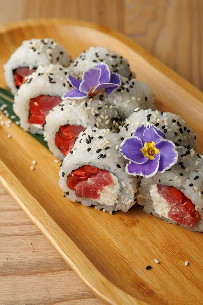 Bambu tabağına yerleştirilmiş balık ve krem peynirli suşi ruloları. Ekmekler çiçekler ve susam tohumlarıyla süslenmiş, basit ama zarif bir sunum sergiliyorlar..