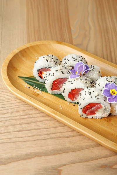 Suşi ruloları taze balıkla doldurulur ve yenilebilir çiçeklerle süslenir ahşap bir tabakta güzelce düzenlenir. Ayarlar yemeğin renkli sunumunu tamamlar.