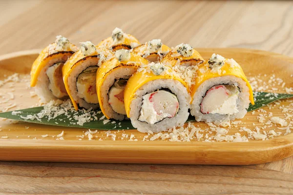Suşi ruloları ahşap bir tabakta güzelce düzenlenir. Ekmeklerde kremalı dolgular ve çıtır çıtır parçalar var. Taze bir yaprak tabakayı süsler, sunumu güçlendirir..