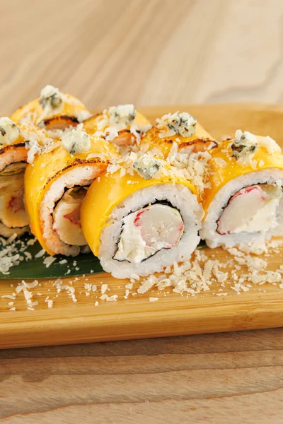 Suşi ruloları ahşap bir tabakta, pirinç katmanları ve dolgular sergileniyor. Üstü sarı sos ve peynirle kaplanmış, lezzetlerin karışımını vurguluyor..