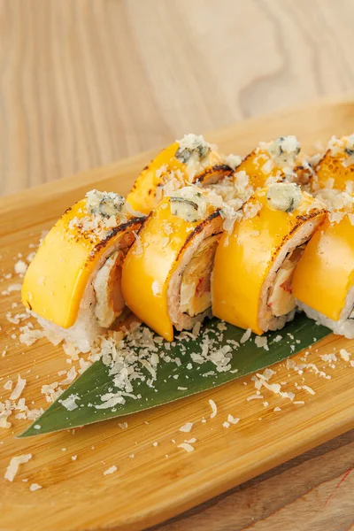 Sarı soslu ve gevreklerle süslenmiş suşi ruloları ahşap bir tabakta servis edilir. Bu yemek, farklı mutfaklardan gelen lezzetlerin karışımını yemek masasında gösteriyor..