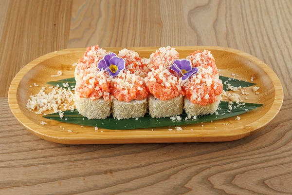 Somon ve ezilmiş malzemelerle kaplı taze suşi ruloları ahşap bir tabakta güzelce düzenlenir. Bir çiçek süslemesi yemeğe biraz renk katar..