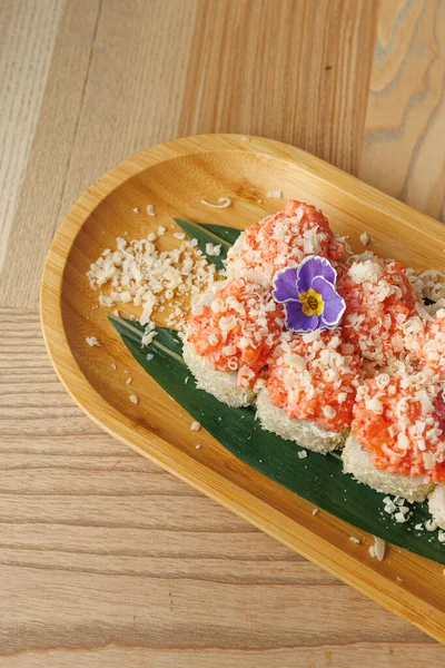 Taze somonla yapılmış lezzetli suşi ruloları gevrek gevrek gevrekler ve yeşil yapraklı çiçek süslemesi. Yemek ortamında ahşap bir tabakta servis edilmiş..