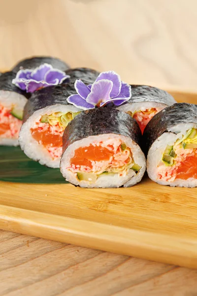 Taze suşi ruloları bambu tabakta sunulur. Ekmekler mor çiçeklerle süslenmiş somon ve sebzelerle dolu. Ahşap yüzey doğal bir dokunuş ekler..