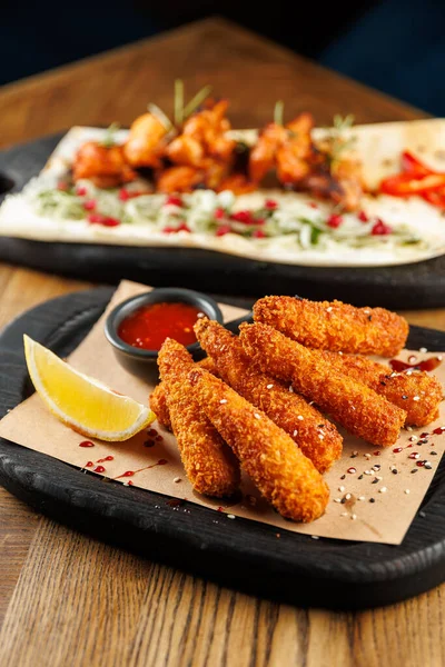 Limon dilimi ve baharatlı sos ile kızartılmış atıştırmalıklar siyah bir ahşap tepsiye konur. Arka planda ızgara tavuk şişleri otlar ve sosla süslenir..