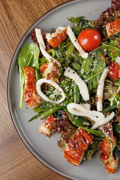 Canlı bir salatada ızgara yılan balığı parçaları, kiraz domatesleri ve susam tohumlu yapraklı yeşillikler bulunur. Tabak, ahşap bir masada basit bir tabakta sunuluyor..