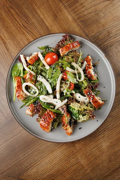 Taze bir salata ızgara tavuk, karışık yeşiller, kiraz domatesleri ve beyaz erişteyle servis edilir. Tabak gri bir tabakta sunuluyor, ahşap bir masaya karşı..