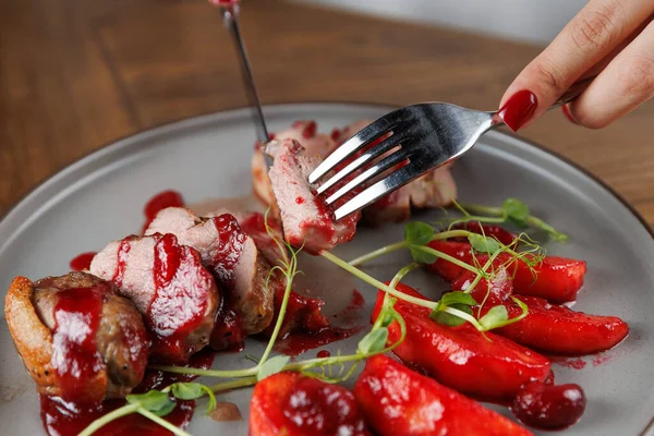 Zengin böğürtlen sosu serpiştirilmiş ördek göğsünü keserken bir el çatal tutuyor. Onun yanında, canlı sebzeler ahşap masadaki tabağa renk katar..