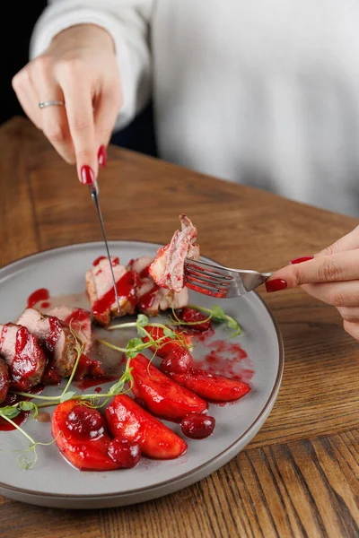 Bir el, ahşap bir masada sunulan böğürtlen sosuyla servis edilen et tabağını keserken bir çatal ve bıçak tutar. Yemek zarif bir şekilde sunuluyor ve canlı renkler sergileniyor..