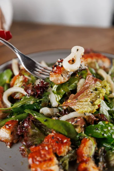 Bir insan bir parça ızgara tavuk ve salatayla çatal tutar. Tabakta yapraklı yeşiller, sebzeler ve bir sos serpintisi yer alıyor. Rahat bir atmosferde öğle yemeği için mükemmel..