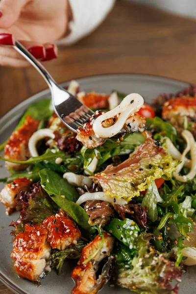 Bir el, bir çatal dolusu renkli salatayı kaldırır. Karışık sebzeler ve ızgara tavukları yuvarlak bir tabakta, ahşap bir masa zeminine yaslar..