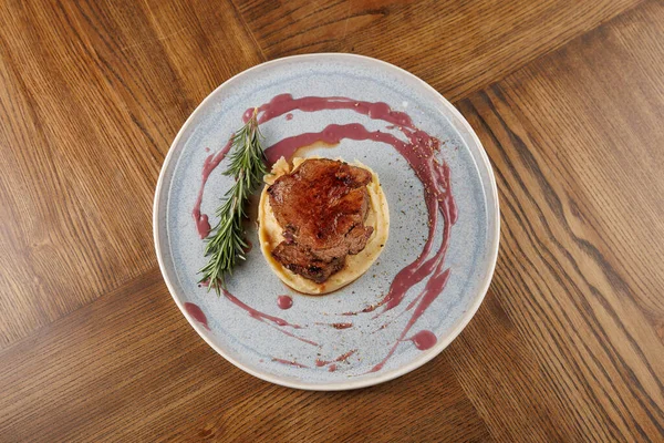 Bir tatlı tabağı, üzerinde et ve sos olan bir hamur işini mavi bir tabakta sergiler. Yanında bir biberiye filizi ile birlikte, bir restoran ortamında ahşap bir masada oturur..