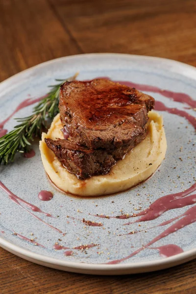 Bir tabak, pürüzsüz patates püresinin üzerinde duran kalın bir et dilimini içeriyor. Tabak biraz sos ve biberiye ile süslenmiş..