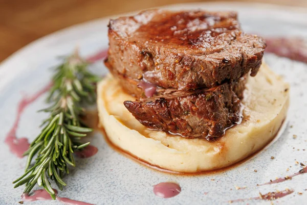 Yumuşak pişirilmiş, yumuşak patates püresinin üzerine yerleştirilmiş, biberiye filizi ile süslenmiş, doyurucu bir biftek. Yemek şık bir tabakta, lezzetleri vurguluyor..