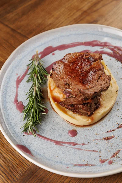 Kremalı patates püresi üzerine ızgara sığır eti. Taze biberiyeyle süslenmiş ve böğürtlen sosuyla sulandırılmış. Tabak ahşap bir masada yuvarlak mavi bir tabakta servis edilir..
