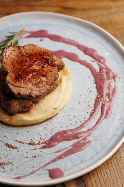 Sığır filetosu pürüzsüz patates püresi yatağında, mavi bir tabakta çilek sosuyla süslenmiş. Kırsal bir ahşap masa yemek için sıcak bir atmosfer sağlar..