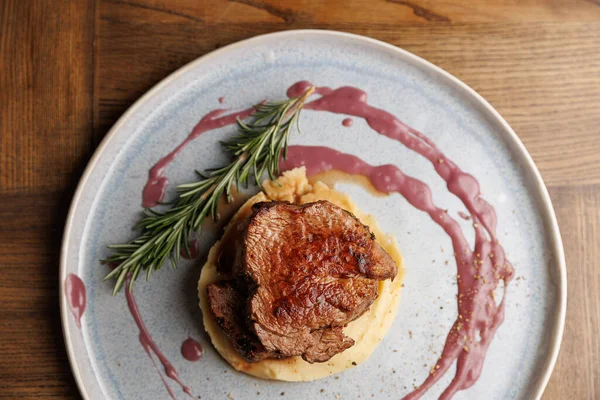 Et tabağı, pürüzsüz bir patates püresi tabakasının üzerinde, üzerine sos serpiştirilmiş ve ahşap bir masada taze biberiye ile süslenmiş. Ayarlar aşçılık sanatını ön plana çıkarıyor.