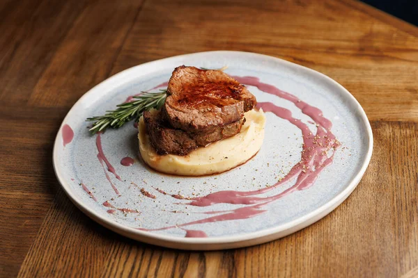 Kurutulmuş et, açık mavi bir tabakta kremalı patates püresinin üzerinde oturur. Bir sos yağmuru tabağı sarar ve biberiye filizi bir tutam renk ve tat ekler..