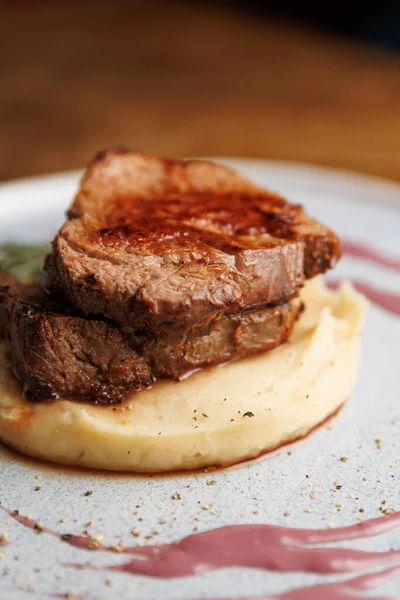 Dilimlenmiş domuz eti, et ve patatesin dokusunu gösteren kremalı patates püresi yatağında servis edilir. Bu yemek bir restoran ortamında aşçılık perspektifini vurgular.