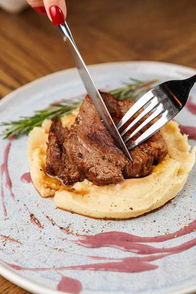 Bir el bıçak ve çatal tutuyor, patates püresinin üzerine yerleştirilmiş lezzetli bir parça eti kesmeye hazır, otlarla süslenmiş, şık bir tabakta..