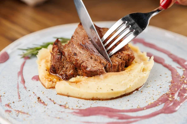 Bir insan bir et parçasını patates püresi ve sos serpiştirilmiş bir tabakla keser. Yeşil otlar taze bir dokunuş katar. Bu yemek sıcak bir yemek atmosferinde keyiflidir..