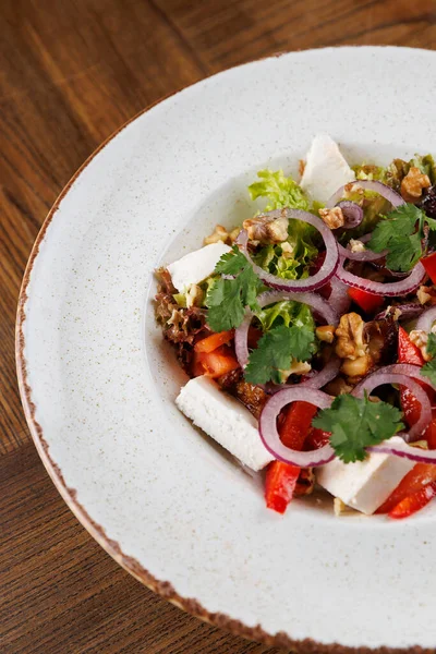 Taze bir salatada kırmızı dolma biber, soğan ve ceviz ve bitkilerle karıştırılmış yeşil marul da dahil olmak üzere doğranmış sebzeler yer alıyor. Tahta bir masada beyaz bir tabakta servis edilmiş..