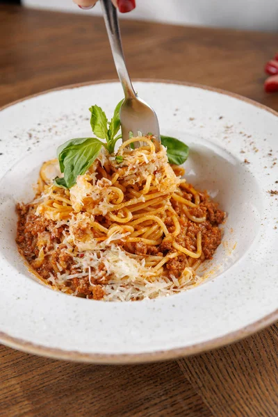 Birisi beyaz tabakta peynirli et soslu makarna döndürmek için çatal kullanıyor. Ahşap masa bir restorandaki yemek deneyimine sıcaklık katar..