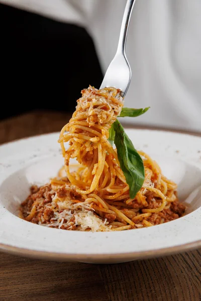 Bir çatal, et sosuyla kaplı spagettiyi beyaz bir kaseden kaldırır. Tepesine yeşil fesleğen konur ve rendelenmiş peynir tabağın üzerine serpilmiştir. Arka planda bir yemek salonu görünüyor..