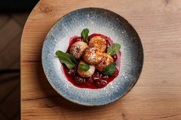 Bir tabakta, üzeri toz şekerle kaplı, kiraz sosuyla çevrili ve ahşap bir yüzeyde taze nane ile süslenmiş yumuşak hamur tatlısı sergileniyor..