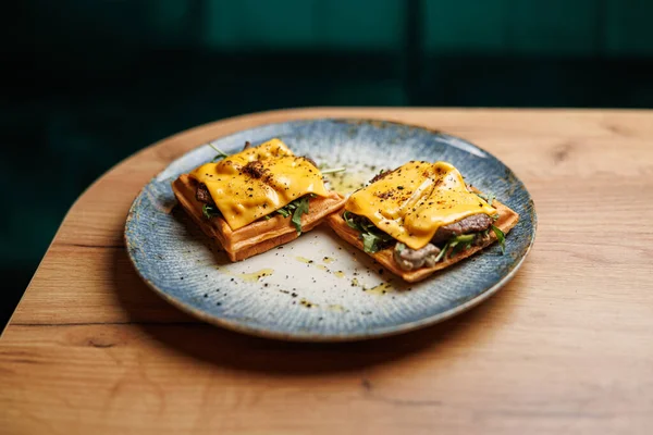 Erimiş peynir, yeşillik ve sığır eti içeren iki açık yüzlü sandviç dekoratif bir mavi tabağa yerleştirilir. Bu tabak ahşap bir masada sıradan bir yemek masasında servis edilir..