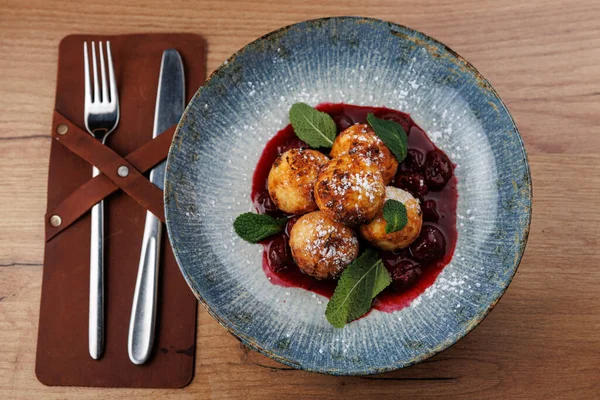 Hamur köfteleri kiraz sosuyla mavi bir tabağa konur ve nane yapraklarıyla süslenir. Yakındaki kahverengi bir peçetenin üzerinde bir çatal ve bıçak duruyor. Bu sahnede çeşitli mutfak stilleri gösteriliyor.