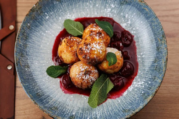 Beş yuvarlak tatlı topu taze nane yapraklarıyla süslenmiş bir böğürtlen sosunun üzerine oturur. Tabak renkli ve davetkar. Mavi bir kase ve ahşap bir masa var..