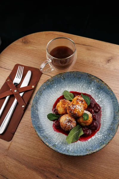 Bir kase tatlı peynirli hamur köftesi nane sosuyla süslenmiş kiraz sosunda oturur. Yakınlarda, bir fincan çay, çatal bıçaklı ahşap bir masanın üzerinde duruyor..