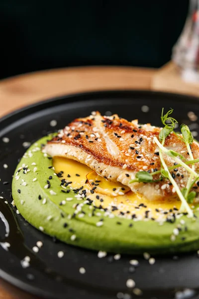 Izgara balık filetosu, susam tohumları ve taze otlarla süslenmiş kremalı yeşil bir sosun üzerinde duruyor. Bu yemek şık bir restoran ortamında lezzet karışımı sergiliyor..