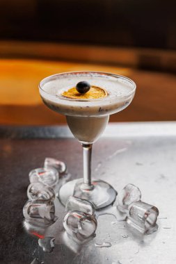 Parlak metal bir yüzey üzerinde bir kokteyl bir bardakta durur. Buz küpleri camın etrafına saçılmış ve içkinin üzerinde bir garnitür duruyor..