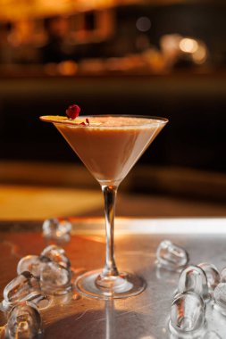 Bir kokteyl, gül yaprağıyla süslenmiş berrak bir bardakta oturur. Üssü çevreleyen buz küpleri ve arka planda sıcak ışıklandırmalı bir bar görülebilir..
