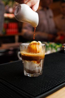 Bir kişi yoğun bir kafede kremalı dondurmayla dolu bir bardağın üzerine zengin bir kahve şurubu döküyor. Hareketli atmosfer yemek hazırlama sahnesine ekleniyor..